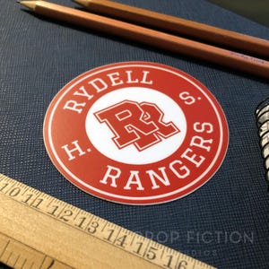 Op de afbeelding: Ronde sticker in rood en wit met de tekst "Rydell H.S. Rangers" en een rood "R" logo.