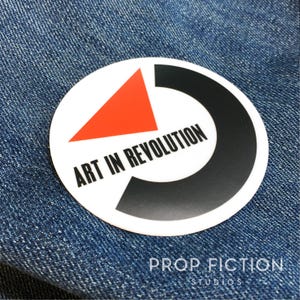 May include: A white circular sticker with a black and red design. The sticker features the text "Art in Revolution" in black lettering.