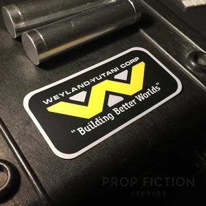 Op de afbeelding: Zwarte sticker met de tekst "WEYLAND-YUTANI CORP" en "Building Better Worlds", met een gele "W" logo. Twee zilveren cilinders zijn zichtbaar. De sticker heeft een witte rand.