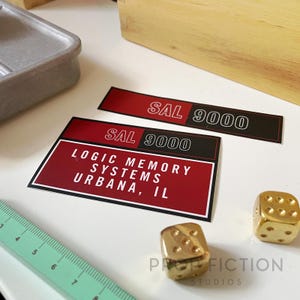 May include: Two gold dice and two red and black SAL 9000 labels are displayed on a white surface. One label reads "LOGIC MEMORY SYSTEMS URBANA, IL." A green ruler is in the lower left corner. The text "PROP FICTION STUDIOS" is at the bottom.