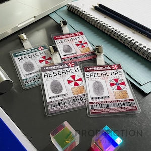 Puede incluir: Cuatro tarjetas de identificación transparentes de Umbrella Corporation con diferentes títulos: Médico, Seguridad, Investigación y Operaciones Especiales. Cada tarjeta presenta una huella dactilar, un logotipo y un código de barras. Las tarjetas están sujetas.