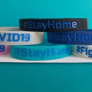 May include: Five colorful silicone wristbands with different messages. The wristbands are white, pink, teal, blue, and black. The messages on the wristbands are "#FightCOVID19", "#StayHome", "COVID19", "#StayHome", and "#StayHome".