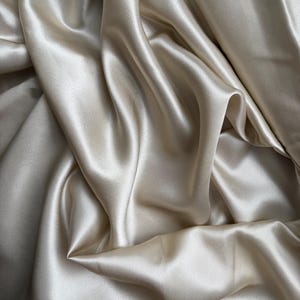 Könnte beinhalten: Ein glatter, glänzender Stoff in hellem Champagner-Beige. Das Material weist eine hochglänzende Oberfläche mit tiefen, fließenden Falten und weichen Schatten auf, die eine dynamische, gewellte Textur über die gesamte Stoffoberfläche erzeugen.