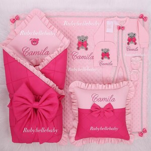 May include: A pink baby bedding set featuring a blanket, swaddle, pillow, hat, and onesie. The set includes a large bow, teddy bear accents, and the name "Camila" embroidered on several items. The brand name "Rubybellebaby" is also visible.