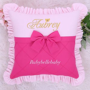 May include: A pink and white decorative pillow with a ruffled edge. The pillow features the name "Aubrey" embroidered in gold script above a pink bow. The lower section is a quilted pink fabric with the text "Rubybellebaby".