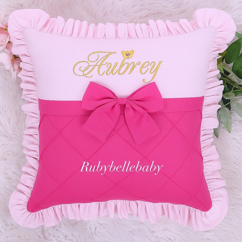 May include: A pink and white decorative pillow with a ruffled edge. The pillow features the name "Aubrey" embroidered in gold script above a pink bow. The lower section is a quilted pink fabric with the text "Rubybellebaby".