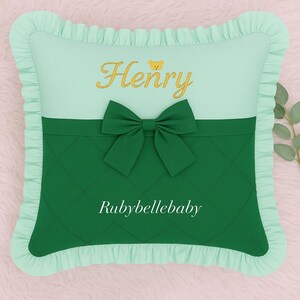 May include: A square, mint green pillow with a ruffled border. The top section is mint green with the name "Henry" embroidered in gold, and a small bear icon. A dark green bow is centered on the pillow, with the text "Rubybellebaby" below.