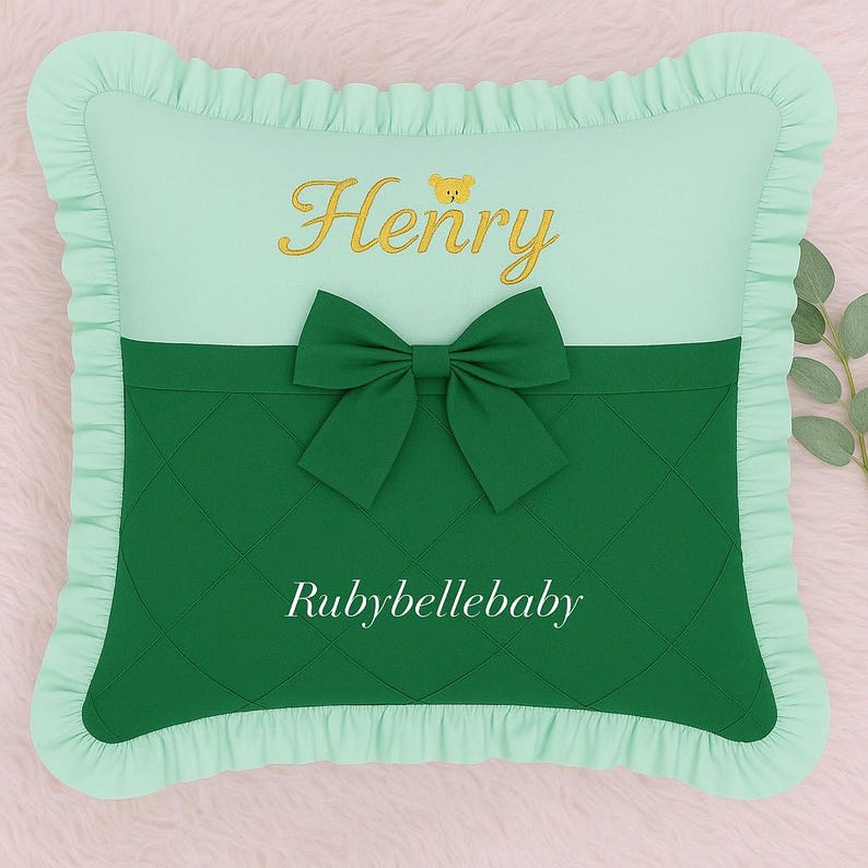May include: A square, mint green pillow with a ruffled border. The top section is mint green with the name "Henry" embroidered in gold, and a small bear icon. A dark green bow is centered on the pillow, with the text "Rubybellebaby" below.