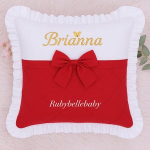 May include: A white pillow with a red quilted section and a red bow. The pillow has the name "Brianna" embroidered in gold, with a small teddy bear icon. The text "Rubybellebaby" is also visible.