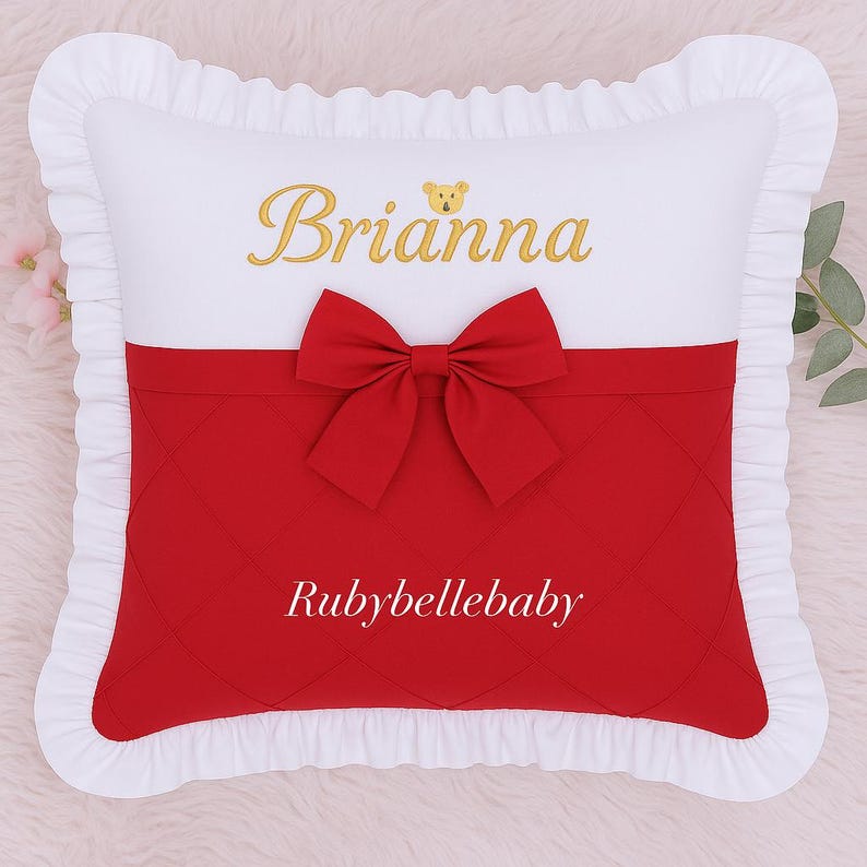 May include: A white pillow with a red quilted section and a red bow. The pillow has the name "Brianna" embroidered in gold, with a small teddy bear icon. The text "Rubybellebaby" is also visible.