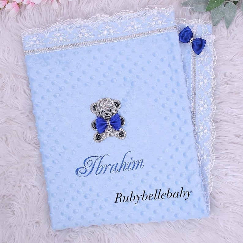 May include: A light blue baby blanket with a textured surface, a white lace trim, and a silver and blue bow. The blanket features a jeweled teddy bear and the name "Ibrahim" embroidered in blue. The text "Rubybellebaby" is also visible.