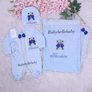 May include: A baby blue newborn set featuring a hat, onesie, mittens, and blanket. Each item is adorned with a teddy bear applique and the name "Ibrahim". The blanket and onesie have the text "Rubybellebaby". The set is trimmed with lace and a blue bow.