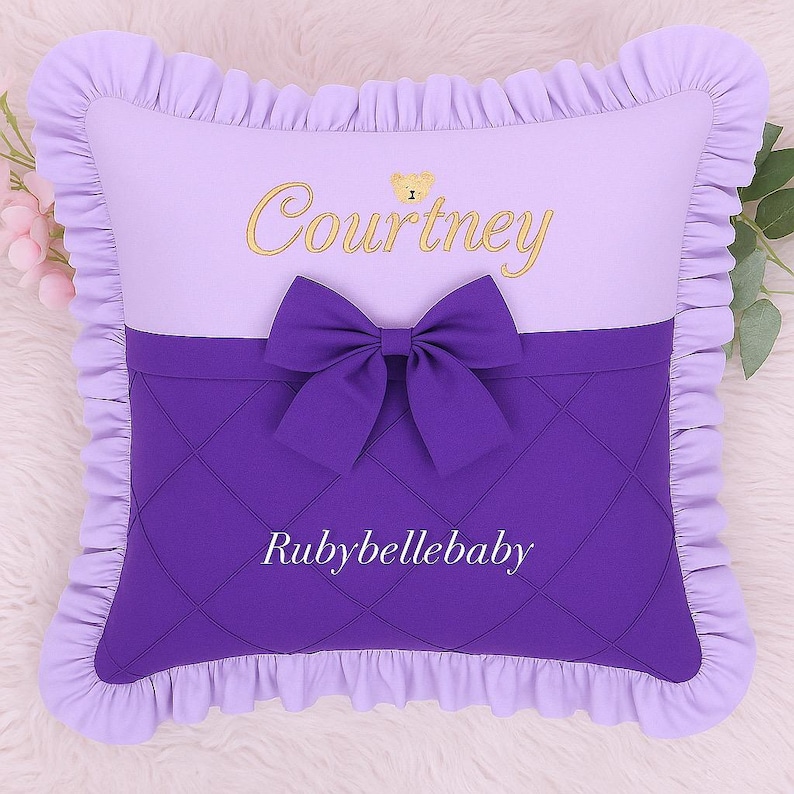 May include: A lavender and purple decorative pillow with a bow. The top half is lavender with the name "Courtney" embroidered in gold. The bottom half is quilted purple with the text "Rubybellebaby".