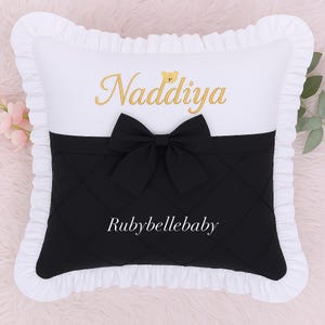 May include: A decorative pillow with a white top and ruffled edges. The pillow features a black quilted bottom section with a black bow. The name "Naddiya" is embroidered in gold script, and the text "Rubybellebaby" is printed in white.