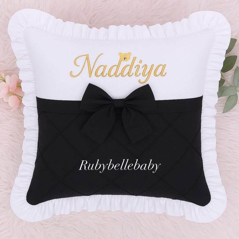 May include: A decorative pillow with a white top and ruffled edges. The pillow features a black quilted bottom section with a black bow. The name "Naddiya" is embroidered in gold script, and the text "Rubybellebaby" is printed in white.