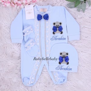 May include: A light blue baby outfit set, including a onesie, hat, and mittens. The onesie features a bow tie, lace trim, and a teddy bear applique. The name "Ibrahim" is embroidered on the onesie and hat. The set is designed for newborns.