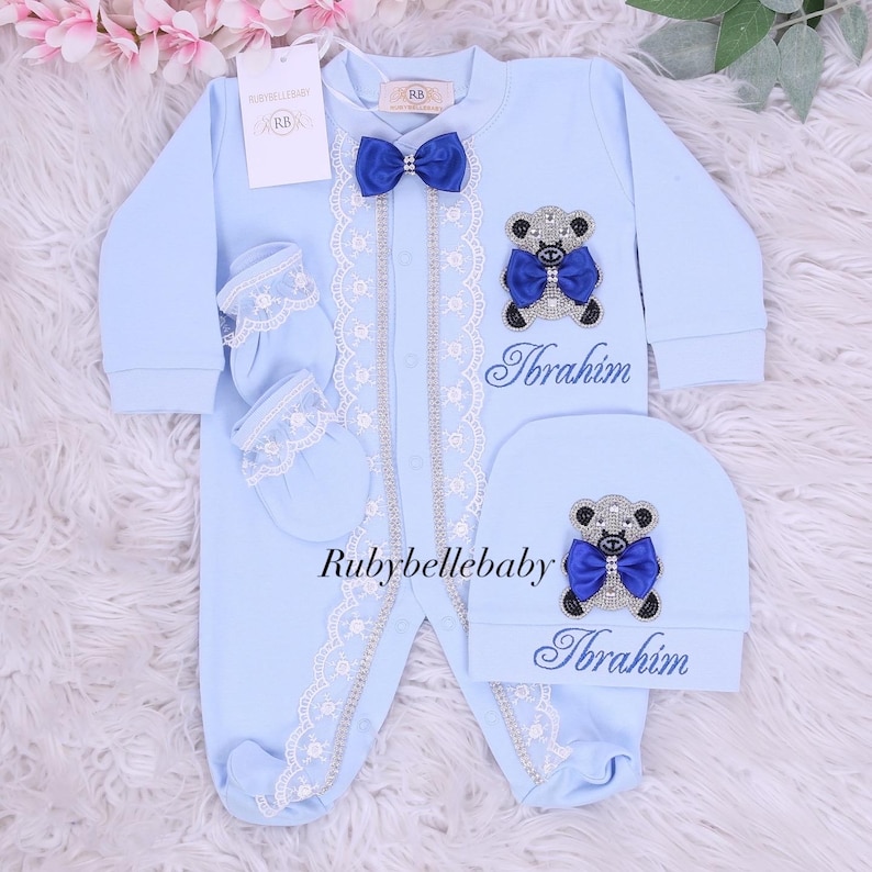May include: A light blue baby outfit set, including a onesie, hat, and mittens. The onesie features a bow tie, lace trim, and a teddy bear applique. The name "Ibrahim" is embroidered on the onesie and hat. The set is designed for newborns.