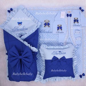 May include: A baby bedding set in shades of blue, featuring a blanket, swaddling wrap, pillow, hat, and onesie. Each item is adorned with a teddy bear design, bows, and the name "Ibrahim". The set includes lace and ruffled edges.