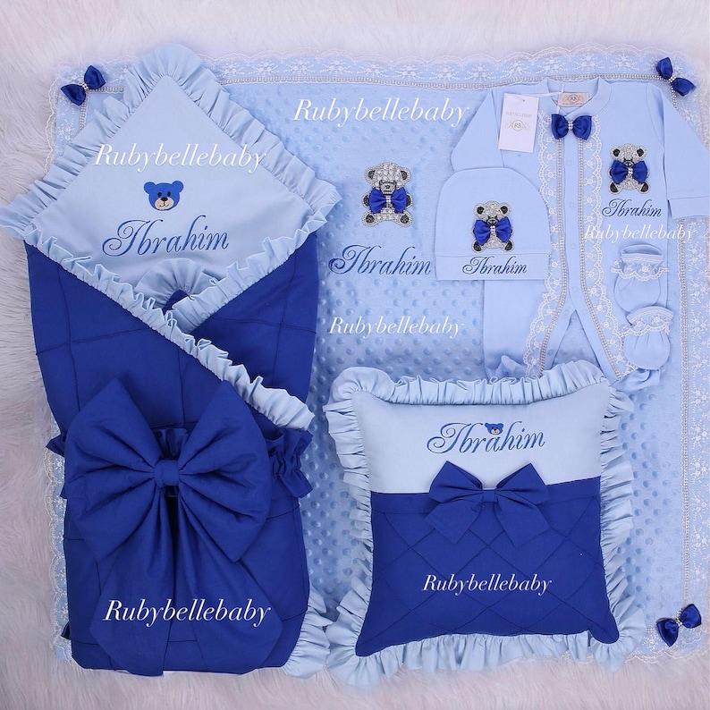 May include: A baby bedding set in shades of blue, featuring a blanket, swaddling wrap, pillow, hat, and onesie. Each item is adorned with a teddy bear design, bows, and the name "Ibrahim". The set includes lace and ruffled edges.