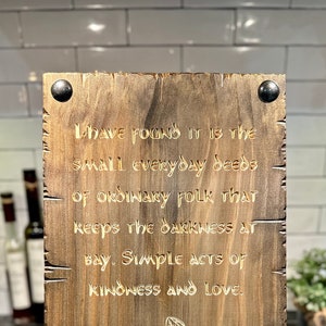 May include: A wooden sign with a quote from Gandalf, a character from the Lord of the Rings. The quote reads: "I have found it is the small everyday deeds of ordinary folk that keeps the darkness at bay. Simple acts of kindness and love."