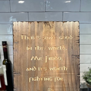 May include: A wooden sign with a quote from the Lord of the Rings: "There's some good in this world, Mr. Frodo, and it's worth fighting for." The quote is carved into the wood and the sign is stained a dark brown. The sign also includes the name "Samwise" at the bottom.