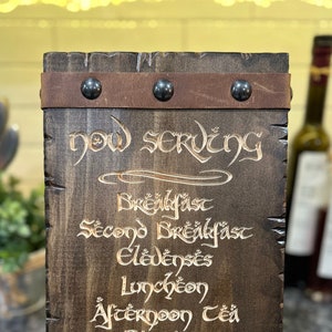 May include: A dark wooden sign with the words "Now Serving" and a list of meal times in an elegant font. The sign is framed with brown leather straps and metal studs, and is designed to stand upright.