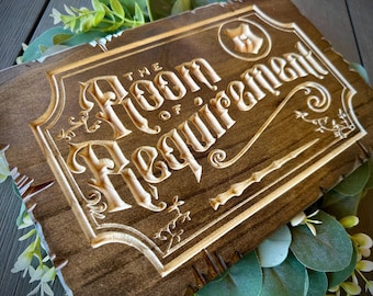 May include: A wooden sign with the text "The Room of Requirement" carved into it. The sign is brown with a light brown wood grain. The text is in a stylized font and is surrounded by decorative flourishes.