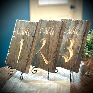 May include: Three rustic wooden table number signs with black metal stands. The signs are distressed brown wood with the words "Table" and the numbers "1", "2", and "3" burned into the wood.