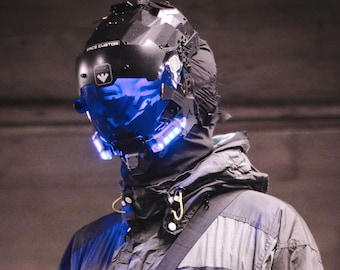 May include: A person wearing a gray jacket with a black bag slung over their shoulder. The person is wearing a black helmet with a blue visor and blue lights. The helmet has the text "FACE CUSTOM" on it. The jacket has the text "XTRMWORKS" on it.