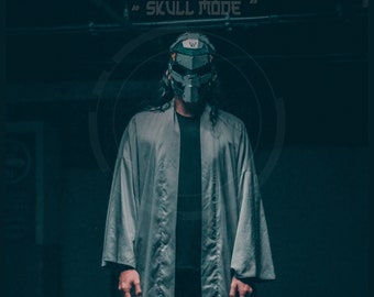 May include: A person wearing a gray robe and a black and silver helmet with the text "SKULL MODE" above. The helmet has a skull design and the text "MASKSHIELD MK.29 HADEs" below.