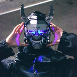 May include: A futuristic, black and gray mask with glowing blue lights. The mask has a horned design and is worn over the face. The image is of a person wearing the mask, with their hands framing the sides of the mask.