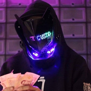 Op de afbeelding: Een persoon draagt een zwarte hoodie met een zwart en blauw LED-masker met de tekst "CUSTOM" erop. De persoon houdt een stapel biljetten in zijn handen.