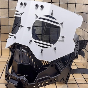 May include: A white and black 3D printed mask with a large, round, open-faced visor. The mask has a black, intricate, geometric design and a strap for securing it to the head.