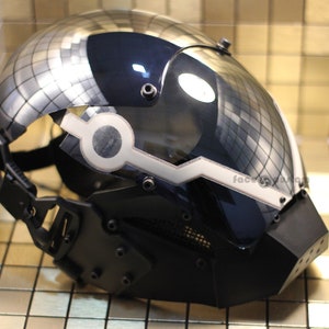 May include: A black and silver helmet with a clear visor and a white stripe across the front. The helmet has a textured surface and a black strap.