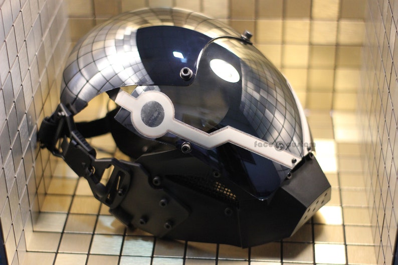 May include: A black and silver helmet with a clear visor and a white stripe across the front. The helmet has a textured surface and a black strap.
