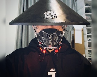 May include: A person wearing a black hooded jacket with a clear plastic face mask and a black hat with a circular design. The mask has red lights on the sides.