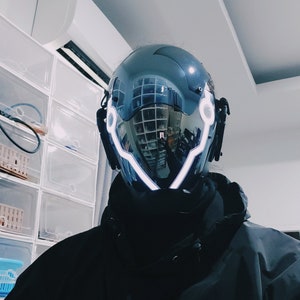 May include: A black helmet with a reflective visor and white LED lights around the edges. The helmet has a futuristic design.