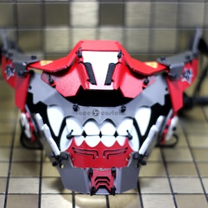 May include: A red and grey face mask with a white and black teeth design. The mask has a geometric design and is made of plastic. The text "face custom" is printed on the mask.