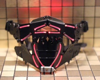 May include: A black mask with red LED lights. The mask has a geometric design and is made of plastic.