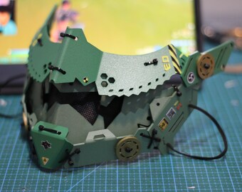 May include: A green and black detailed mask with a textured surface and black straps. The mask has a geometric design with various shapes and cutouts. The mask has a yellow and black warning label with the text "D3".