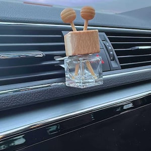 May include: A car air freshener featuring a clear, square glass bottle and a wooden top. Two wooden lollipop-shaped diffusers are attached to the top. The air freshener is placed on a car's dashboard air vent.