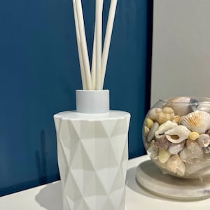 May include: White ceramic diffuser bottle with a geometric pattern and white diffuser sticks. The bottle is on a white surface next to a glass bowl filled with seashells.