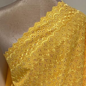 May include: A close-up of a yellow eyelet fabric draped over a mannequin. The fabric features an intricate floral and swirl pattern with a scalloped edge. The eyelet fabric is a bright, sunny yellow.