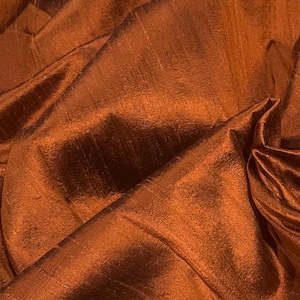 Puede incluir: Primer plano de una tela texturizada de color cobre con un brillo sutil. El material está doblado y drapeado, creando sombras y reflejos que enfatizan su rico color y textura superficial. La tela parece ser un textil lujoso.