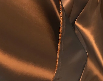 Op de afbeelding: Close-up van een bruine satijnen stof met een licht getextureerd oppervlak. De stof is dubbelgevouwen, waardoor de rauwe rand zichtbaar is.