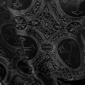May include: Close-up of a black fabric with an ornate, repeating pattern. The design features crosses, floral elements, and swirling details in a lighter shade of black, creating a textured, embossed effect. The fabric appears to be draped.
