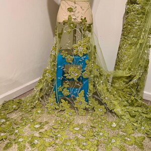 May include: Lime green floral embroidered fabric. The sheer material features large, three-dimensional flowers with beaded accents. The fabric is draped over a mannequin and a rolled section of the fabric is visible on the right side.
