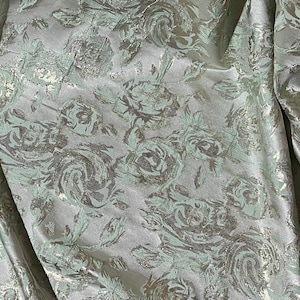May include: Silver fabric with a raised, embossed pattern of green roses. The fabric has a subtle sheen and a soft, luxurious feel.