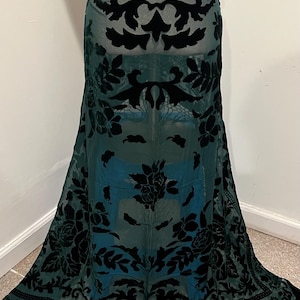 Puede incluir: Falda larga de color verde azulado oscuro con un patrón floral de terciopelo negro. La falda tiene una cintura alta, una superposición transparente y un dobladillo con flecos. El diseño floral incluye rosas y otras flores estilizadas.
