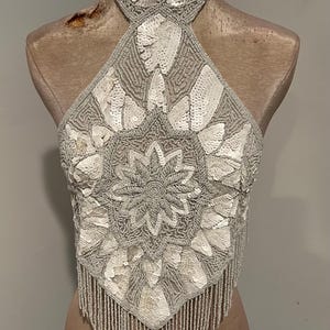 Puede incluir: Top halter plateado con cuello choker de cuentas y dobladillo con flecos. El top está adornado con lentejuelas y cuentas blancas, formando un patrón floral en la parte delantera. Se muestra en un maniquí.